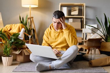 Sarı kapüşonlu stresli bir adam dizüstü bilgisayarı üzerinde bağdaş kurarak oturur ve endişeli bir ifadeyle kutuları ve bitkileri taşır..
