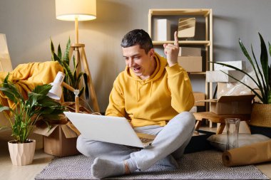 Bir adam yeni evinde dizüstü bilgisayarını kullanırken heyecanlı ve yukarı bakan kutuların arasında oturuyor..