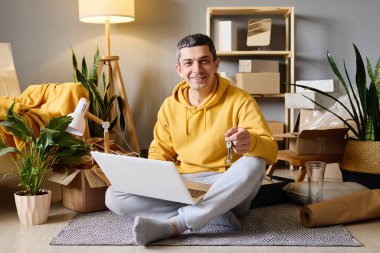 Gülümseyen bir adam dizüstü bilgisayarı ve yeni evinin anahtarlarıyla yeni evindeki kutuları taşırken yerde oturuyor..