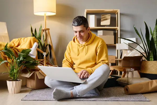 Bir adam yeni evine taşındıktan sonra dizüstü bilgisayarını kullanarak kutuları taşırken yerde oturuyor..