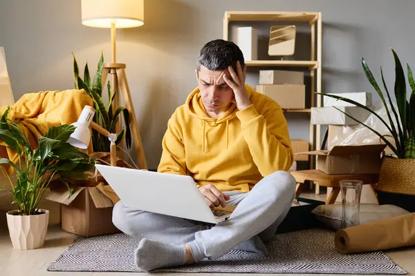 Sarı kapüşonlu stresli bir adam dizüstü bilgisayarı üzerinde bağdaş kurarak oturur ve endişeli bir ifadeyle kutuları ve bitkileri taşır..