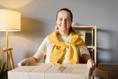 Omuzlarının üzerinden sarkan sarı kazaklı, gülümseyen bir kadın, izleyiciye doğru bakan bir karton kutuyu tutuyor..