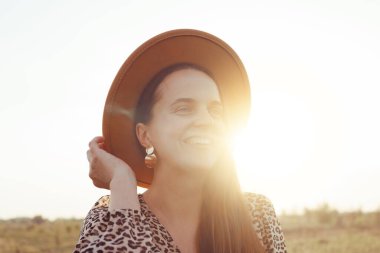 Gülümseyen bir kadın leopar desenli bir elbise, kahverengi bir şapka ve altın küpeler giyiyor..