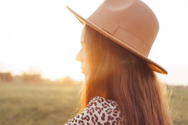 Geniş kenarlı şapka ve leopar desenli elbise giyen bir kadının günbatımına doğru baktığı profil görüntüsü..