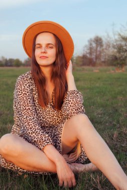 Kahverengi şapkalı ve leopar desenli elbisesi olan bir kadın tarlada çimlerin üzerinde oturmuş kameraya bakıyor..