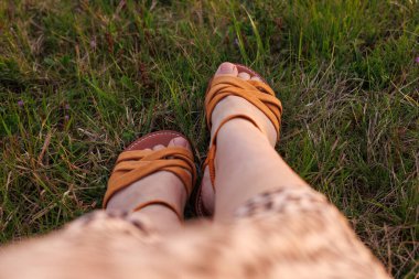 Yeşil çimlerde kahverengi kayışlı sandaletler giyen ayaklara bakan birinci şahıs bakış açısı. Kalıplı bir elbisenin kenarı görünür..