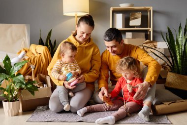 Dört kişilik bir aile yeni evlerinde oyuncaklarla oynuyor. Etrafı kutularla ve ev bitkileriyle çevrili..
