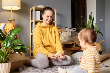 Sarı kapüşonlu, gülümseyen genç bir kadın yerde oturuyor, oyuncağı taşınan kutuların arasında oturan bir bebeğe gösteriyor..
