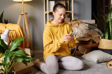 Bir kadın, kutuları ve bitkileri taşıyan bir odada akıllı telefonunu kullanırken gülümser..