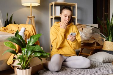 Şaşırmış bir kadın, taşınan kutular ve bitkilerle dolu bir odada akıllı telefonuna bakar..