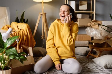 Bir kadın, yeni evine taşındıktan sonra kutuları taşırken telefonda gülerek yerde oturuyor..