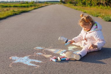 Küçük bir çocuk asfalt bir yolda güneşli bir kırsal bölgede renkli tebeşirli bir aile çiziyor..