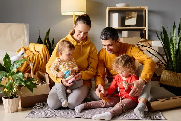 Dört kişilik bir aile yeni evlerinde oyuncaklarla oynuyor. Etrafı kutularla ve ev bitkileriyle çevrili..