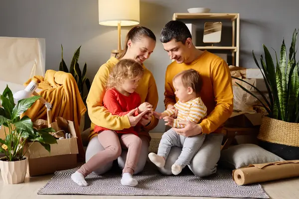 Bir anne ve baba, yeni evlerindeki kutular ve bitkiler arasında sevgi dolu bir şekilde iki küçük çocuğuyla etkileşim halindedir..