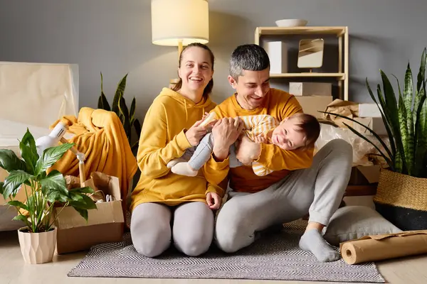 Bebekli mutlu genç bir aile yeni evlerinde kutularını boşaltıp oynuyorlar, gülümsüyorlar ve etkileşime geçiyorlar..
