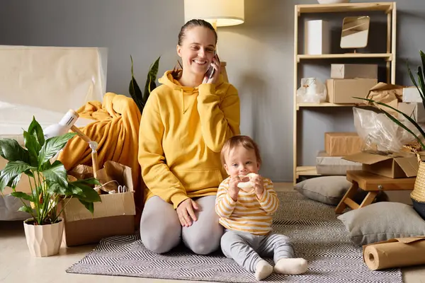 Bir anne, bebeğinin yeni evindeki paketsiz kutuları taşırken telefonuyla konuşurken gülümser..