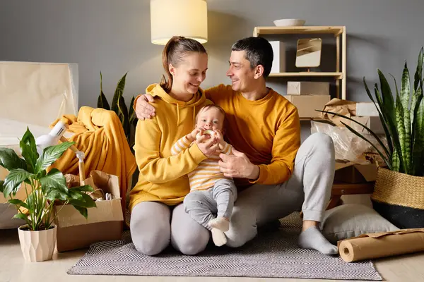 Bebekli, sevgi dolu genç bir aile yeni evlerindeki paketlenmemiş kutuların arasında oturup birbirlerine bakıyorlar..