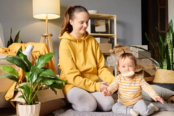 Sarı kapüşonlu genç bir kadın paketleri boşaltılmış kutular ve bitkilerin arasında bir halının üzerinde oturan bebeğe gülümsüyor..