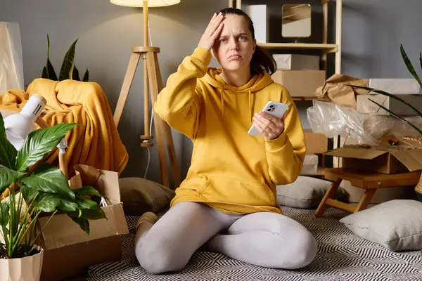 Bir kadın hareket halindeki kutuların arasında oturuyor, stresli görünüyor ve hareket ettikten sonra telefonuna bakarken alnını tutuyor..