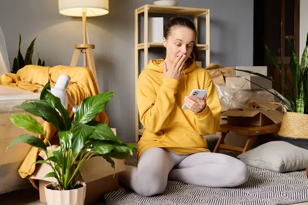 Şaşırmış bir kadın, taşınan kutular ve bitkilerle dolu bir odada akıllı telefonuna bakar..