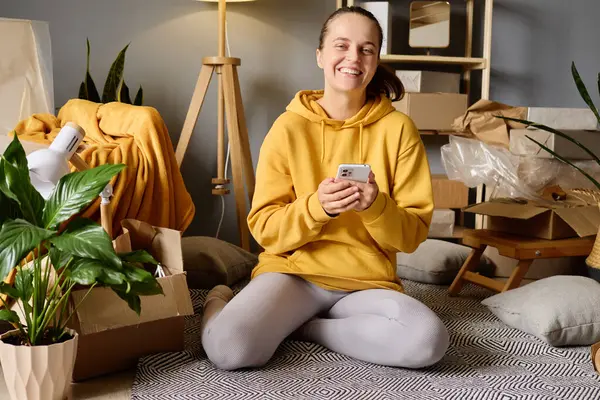 Sarı kapüşonlu, gülümseyen bir kadın bacak bacak üstüne atarak yerde oturuyor. Etrafı kutularla çevrili, elindeki akıllı telefona bakıyor..
