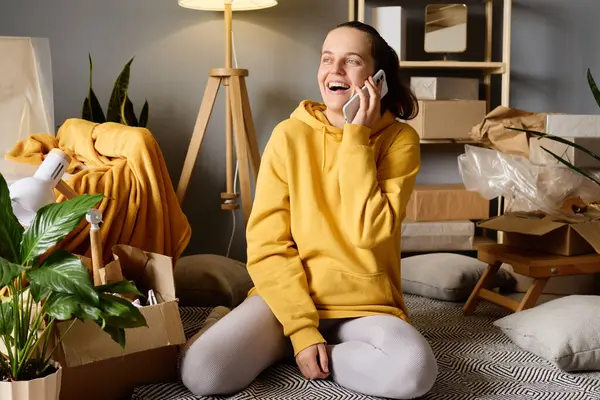 Bir kadın, yeni evine taşındıktan sonra kutuları taşırken telefonda gülerek yerde oturuyor..