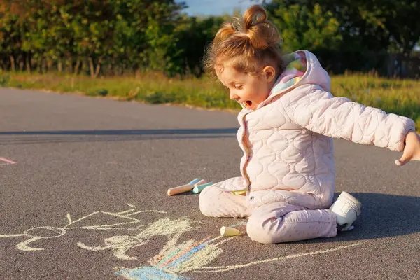 Açık pembe kar kıyafetli küçük bir kız asfalta oturmuş renkli tebeşirle resim çiziyor..