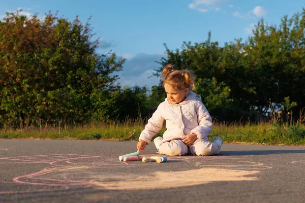 Sarı saçlı, at kuyruklu bir çocuk açık havada asfalta oturmuş güneşli bir günde renkli tebeşirle resim çiziyor..