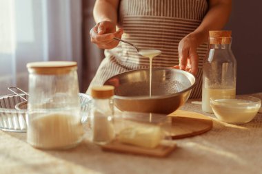 Çizgili bir önlük giyen biri, bir kaşıktan metal bir kaseye kremalı bir sıvı döker ve masada pişirme malzemeleri bulunur..