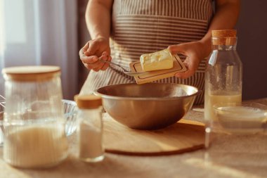 Çizgili önlüklü bir insan, küçük bir ahşap tahtadan tereyağı ekler. Masadaki diğer pişirme malzemeleriyle birlikte metal bir karıştırma kabına..