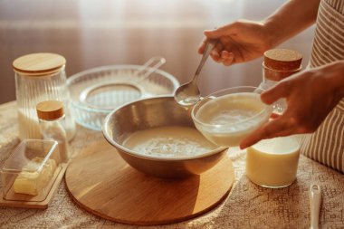 Çizgili önlüklü bir insan, küçük bir cam kaseden daha büyük bir metal kaseye kremalı bir karışım ekler, bir masada pişirme malzemeleriyle çevrili bir kaşıkla..