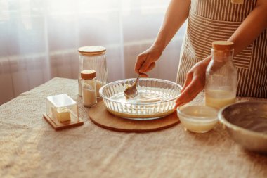 Çizgili önlüklü bir aşçı, cam bir turta tabağına hamur sürer, kaşıkla, pencere kenarındaki bir masada malzemelerle çevrili..