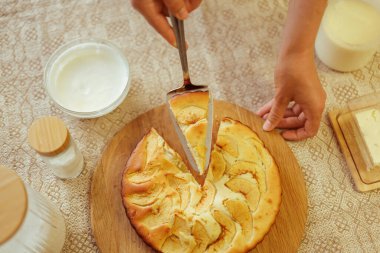 Yukarıdan bakıldığında, ahşap bir tahtanın üzerindeki taze pişmiş elmalı turtadan bir dilim koparmak için spatula kullanan bir aşçı görülüyor. Yanında da bir kase krema ve diğer malzemeler var..