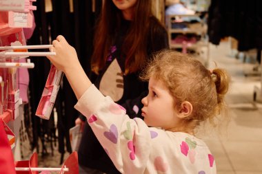 Genç bir kız neşeli bir mağazada renkli güzellikleri incelerken arkadaşı neşeli bir tavırla ona bakıyor..