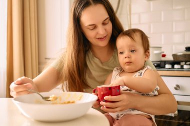 Şefkatli bir anne mutfak masasında oturur ve yemek saatinde bebekle konuşuyormuş gibi görünen bir bebeği kucağında tutar..