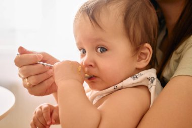 Yakın plan çekimde geniş mavi gözlü bir bebek, kaşıkla besleniyor, kaşığı kendi eliyle tutarken, hassas bir yemek zamanında..