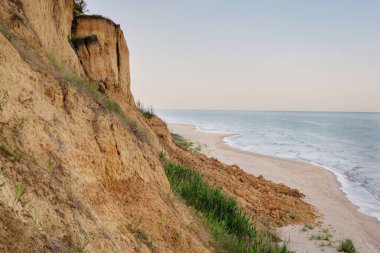 Kumlu bir sahil, uzun, erozyona eğilimli bir uçurum boyunca kıvrılır. Yumuşak, berrak bir gökyüzünün altında dingin bir denizin yumuşak dalgalarıyla karşılaşır..