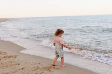 Tulumlu kıvırcık saçlı genç bir çocuk, denizin kenarındaki kumlu bir sahil boyunca yürüyor, yumuşak dalgaların tadını çıkarıyor..