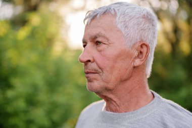 Gri saçlı yaşlı adam, bulanık yeşil bir arka plana karşı düşünceli bir şekilde yana bakıyor..