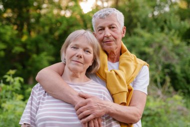Kıdemli çift kameraya sarılıyor ve nazikçe gülümsüyor, yeşil doğayla çevrili..