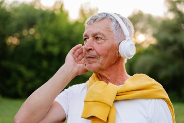 Düşünceli bir son sınıf öğrencisi beyaz kulaklıklardan müzik dinler. Arka planda ağaçlar olan açık havaya bakar..