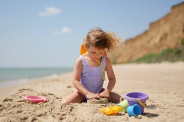 Mayolu ve şapkalı küçük bir kız güneşli bir sahilde renkli oyuncaklarla kumdan bir kale inşa etmeye odaklanıyor..
