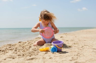 Mor mayo giymiş bir çocuk kumlu bir plajda oturmuş pembe bir eleğe bakarken renkli kum oyuncaklarıyla oynuyor. Arka planda deniz var..
