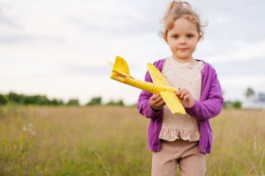 Kıvırcık saçlı tatlı bir kız, bulutlu bir günde çimenli bir tarlada dikilirken doğrudan kameraya bakan sarı oyuncak bir uçak tutuyor..