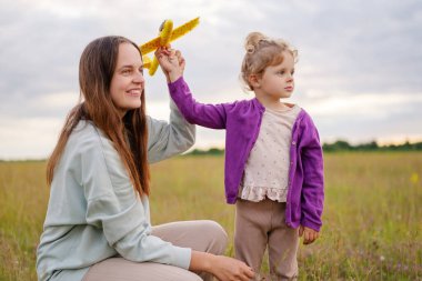 Bir anne ve küçük kızı gün batımında sarı oyuncak bir uçakla oynuyorlar, neşeli bir anı paylaşıyorlar..