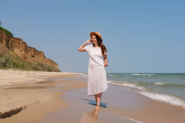 Beyaz elbiseli ve şapkalı bir kadın kayalıkların yakınındaki kumlu bir sahilde yürüyor, deniz kenarında huzurlu bir yaz gününün tadını çıkarıyor..