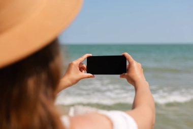 Güneşli bir sahil gününde deniz telefonuyla fotoğraf çeken bir kadın, telefon ve okyanus manzarasına odaklanmış bir şekilde arkadan görülüyor..