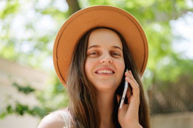 Kahverengi şapkalı neşeli genç bir kadın açık havada akıllı telefonuyla konuşurken gülümsüyor, yumuşak yeşil bir bokeh arka planı var..