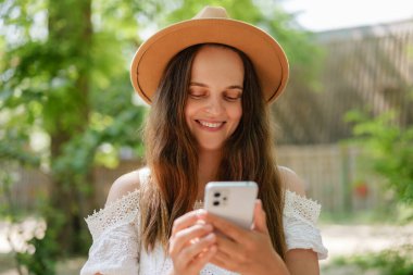 Kahverengi şapkalı ve beyaz dantelli gülümseyen genç bir kadın açık havada telefonuna bakıyor. Etrafı yeşilliklerle çevrili bu güneşli, samimi anda neşe ve sükunet saçıyor..