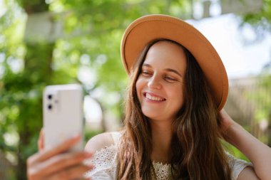 Şık şapkalı genç bir kadın dışarıda selfie çekerken gülümsüyor. Uzun saçları, rahat duruşu ve güneşli arka planı neşeli ve yaz havası yaratıyor..